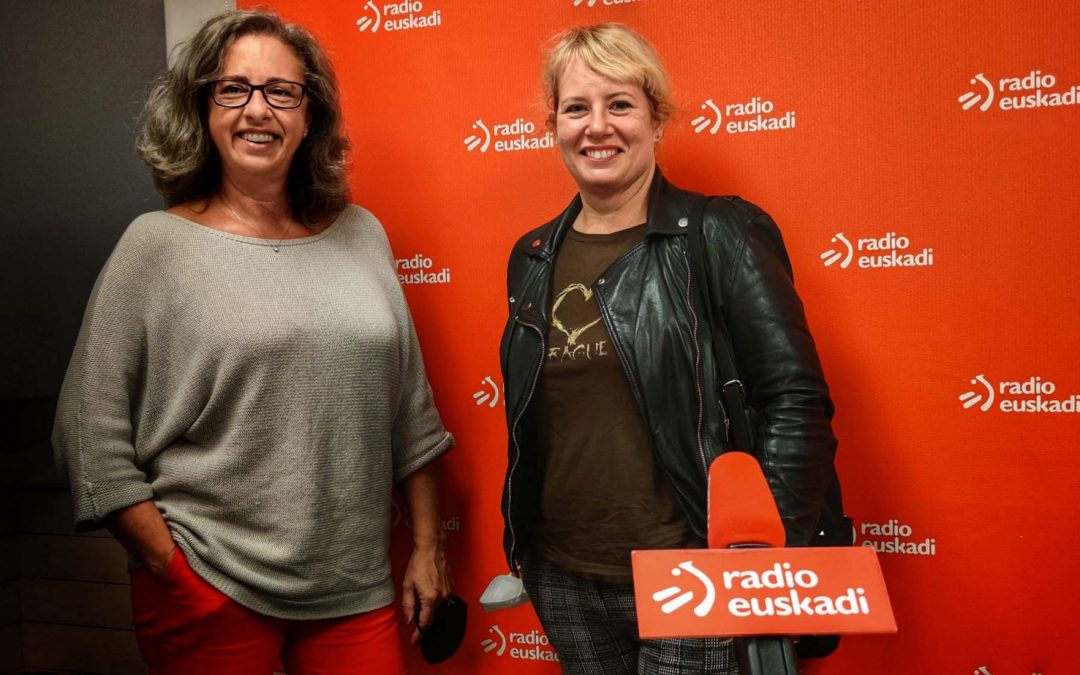 Estudio de Grabación de Radio Euskadi, se puede ver el fondo rojo del color de la emisora y el logotipo del mismo con el texto Radio Euskadi. Aparecen Elixaebte Legarda, vestida de blancco y Arantza Otaduy vestida de marrón y negro, posandofrente al micrófono de la emisora